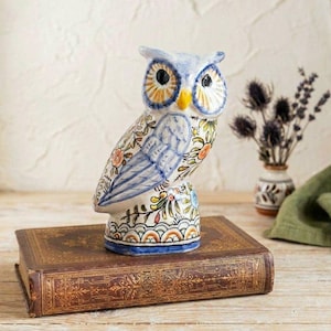 May include: A ceramic owl figurine featuring a hand-painted floral pattern in blue, orange, and green. The owl has large black eyes, a yellow beak, and a base decorated with a scalloped design, standing upright on a rectangular book.