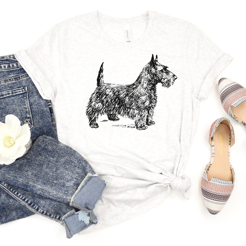 May include: A white t-shirt with a black and white line drawing of a Scottish Terrier dog. The dog is standing on all four legs and looking to the right. The t-shirt is on a white background.