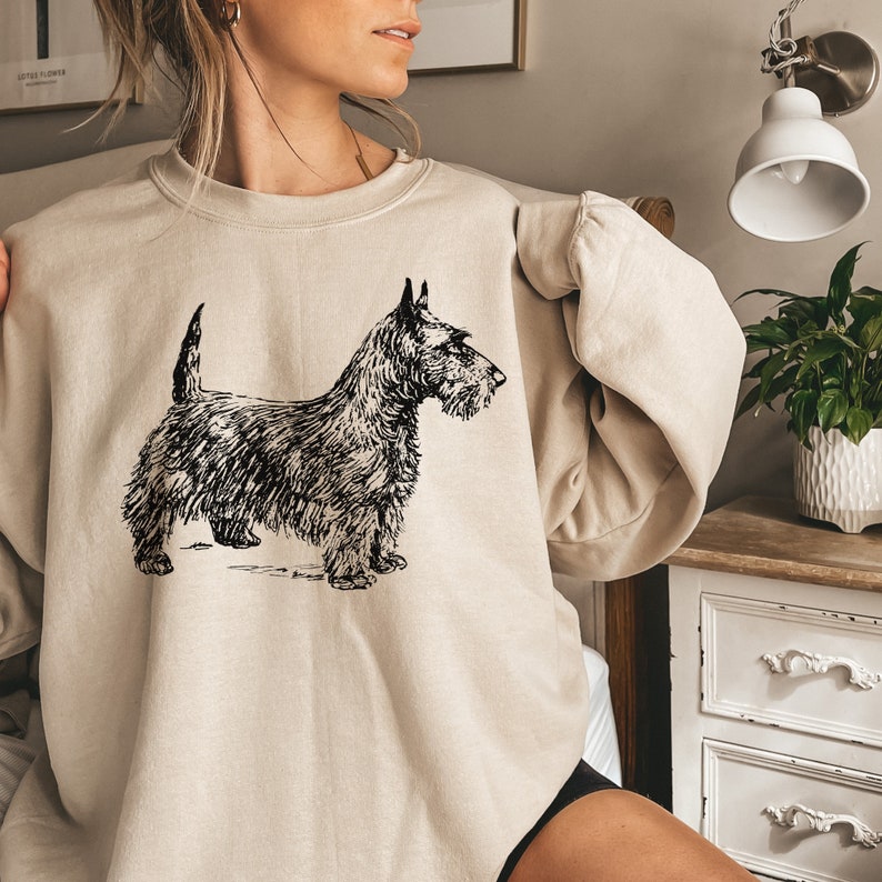 May include: A beige sweatshirt with a black and white illustration of a Scottish Terrier dog.
