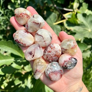 Op de afbeelding: Een verzameling van negen getrommelde stenen met witte, rode en grijze markeringen. De stenen zijn glad en gepolijst en hebben verschillende vormen en maten.