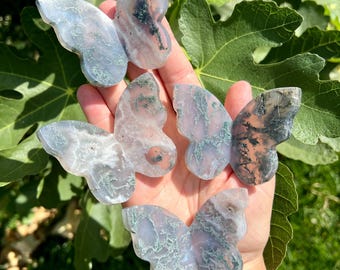 May include: Four butterfly-shaped carvings made of translucent moss agate stone. Each piece features unique dendritic inclusions in shades of dark green, grey, and brown, creating intricate, fern-like patterns within the polished, semi-precious mineral.