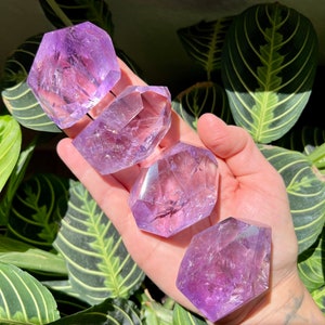 Puede incluir: Cuatro cristales de amatista, cada uno con una forma y un tamaño únicos, se muestran en una mano. Los cristales son de color morado intenso y tienen una textura natural y rugosa.