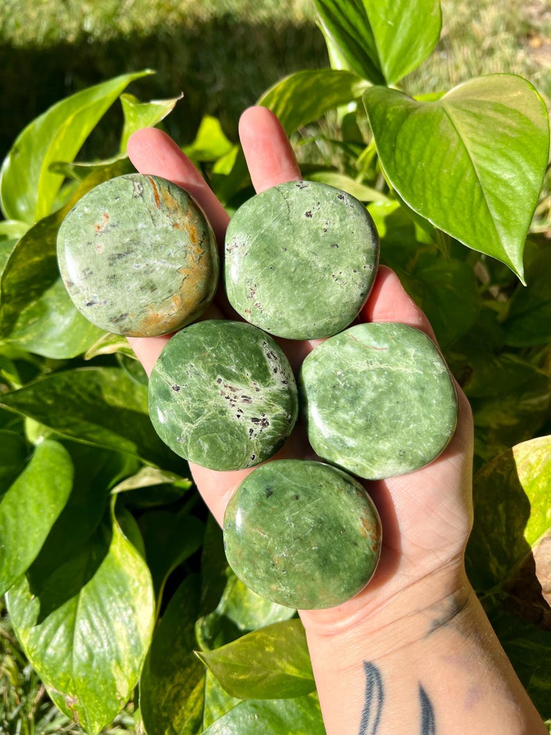 May include: Five polished green stones of varying shapes and sizes are held in a hand. The stones have a mottled appearance with darker green and brown inclusions. The background features lush green foliage.