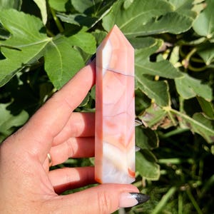 May include: A polished, hexagonal crystal tower featuring a smooth surface with marbled patterns in shades of soft peach, creamy white, and translucent orange, tapering to a pointed tip at the top end of the stone structure.