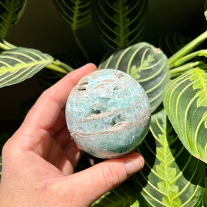 May include: A green and white agate sphere with a swirling pattern. The sphere is smooth and polished, and it is being held in a hand.