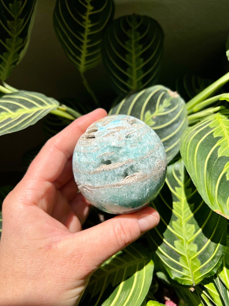 May include: A green and white agate sphere with a swirling pattern. The sphere is smooth and polished, and it is being held in a hand.