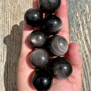 May include: A collection of eight black obsidian spheres with silver streaks. The spheres are smooth and polished, and they are arranged in a pile.