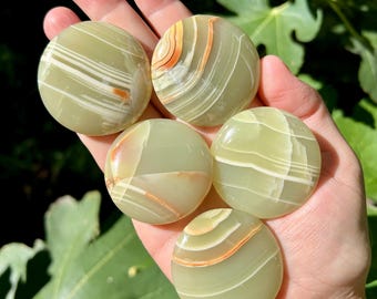 May include: Five round, polished green onyx stones with white and orange banding are held in a hand. The stones have a smooth, glossy surface and vary slightly in their color patterns. The background is blurred with green foliage.
