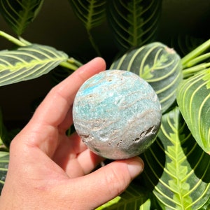 May include: A smooth, round, green and white agate sphere with a swirling pattern. The sphere is being held in a hand.