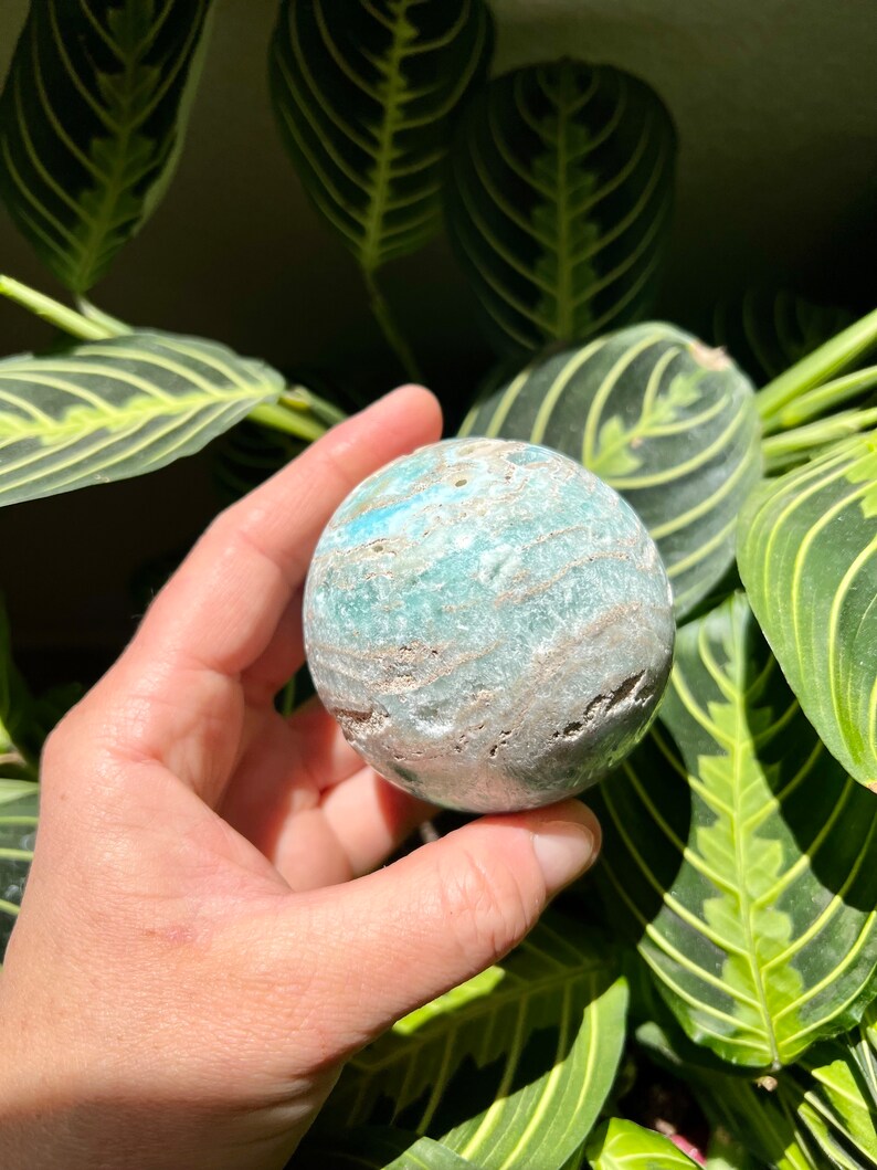 May include: A smooth, round, green and white agate sphere with a swirling pattern. The sphere is being held in a hand.