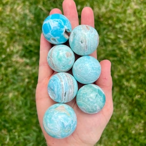 May include: Seven blue and green celestite spheres, also known as celestine, in various sizes and patterns, are held in a hand. The spheres are smooth and polished.