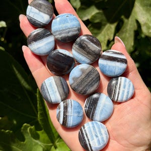 May include: A collection of eleven small, circular, polished stones featuring horizontal bands of light blue, white, and dark charcoal gray. Each stone has a smooth, rounded surface with distinct, layered mineral patterns and a glossy, reflective finish.