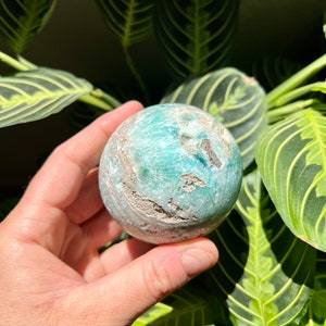 May include: A green and white sphere of polished amazonite, a type of feldspar, with a swirling pattern. The sphere is being held in a hand.