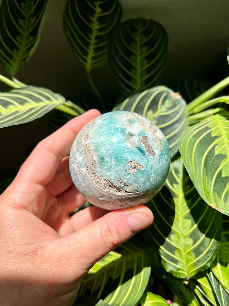 May include: A green and white sphere of polished amazonite, a type of feldspar, with a swirling pattern. The sphere is being held in a hand.