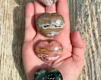 May include: Four heart-shaped polished stones in various colours and patterns. The stones are arranged vertically in a hand, from top to bottom: pink and grey, brown and white, pink and brown, and green with white flecks.