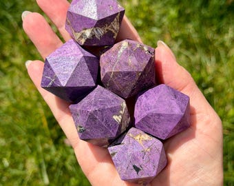 May include: A collection of faceted purple crystals with geometric shapes. The crystals have a deep violet hue with natural variations in color and texture. The crystals are held in a hand against a backdrop of green grass.