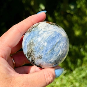 May include: A polished spherical stone featuring a blend of light blue, white, and dark grey mineral inclusions. The surface of the sphere displays a fibrous, chatoyant texture with varying shades of blue and white streaks throughout the round form.
