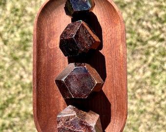 May include: Four dark red garnet crystals, each with a unique shape, are displayed on a wooden tray. The crystals are arranged in a stack, with the largest at the bottom and the smallest at the top.