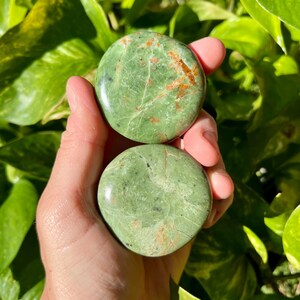 May include: Two round, polished green stones with brown and white veining are held in a hand. The stones have a smooth, glossy surface and are set against a backdrop of green foliage.