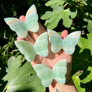 May include: Four carved butterfly figurines in shades of green and tan are displayed against a backdrop of green leaves. The butterfly wings have a striped pattern. The figurines are held in a hand.