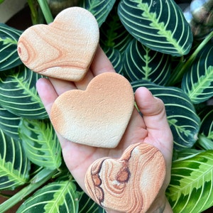 May include: Three heart-shaped sandstone coasters with different patterns. The coasters are a light brown color with red and white streaks.