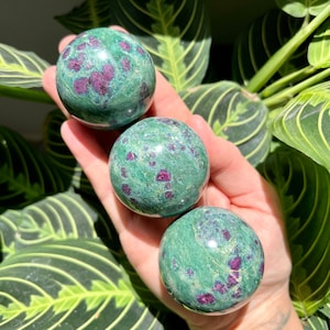 Könnte beinhalten: Drei grüne und rote Rubinzoisit-Kugeln, auch bekannt als Rubin in Zoisit, werden in einer Hand gezeigt. Die Kugeln sind glatt und poliert.