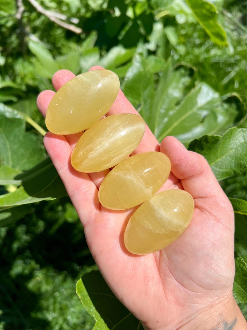 May include: Four yellow calcite tumbled stones, each with a smooth, oval shape. The stones are arranged in a hand, with the largest stone at the top and the smallest stone at the bottom.
