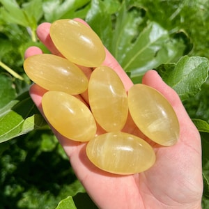 May include: Six yellow tumbled stones, also known as yellow calcite, are arranged in a hand. The stones are smooth and polished, with a light yellow color.