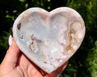 May include: A heart-shaped bowl carved from polished stone featuring a blend of soft pink, lavender, and grey hues. The surface displays natural mineral inclusions, crystalline formations, and swirling patterns throughout the smooth, concave interior.