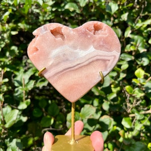 May include: A pink and white heart-shaped agate crystal on a gold metal stand. The crystal has a natural, rough texture and is translucent.