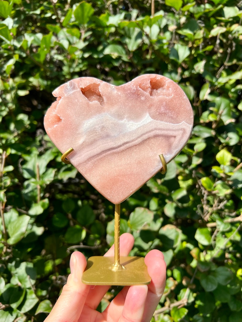 May include: A pink and white heart-shaped agate crystal on a gold metal stand. The crystal has a natural, rough texture and is translucent.
