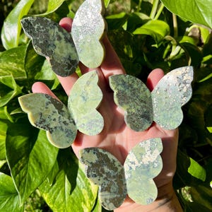 May include: Five green and gray stone butterfly figurines are displayed against a backdrop of green leaves. The butterfly wings have a mottled pattern, with variations in color and texture. The figurines are held in a hand.