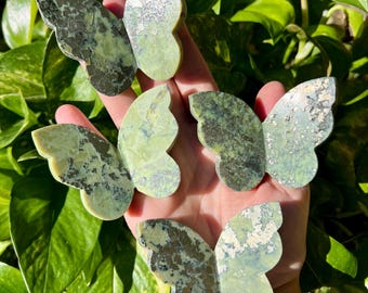 May include: Five green and gray stone butterfly figurines are displayed against a backdrop of green leaves. The butterfly wings have a mottled pattern, with variations in color and texture. The figurines are held in a hand.