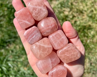 May include: A collection of eight tumbled pink selenite stones. The stones are smooth and polished, with a translucent, milky white appearance. The stones are arranged in a hand, with the largest stone at the top.