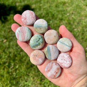 May include: A hand holding a collection of smooth, round stones. The stones vary in color, including pink, green, and beige, with natural patterns and textures. The stones are approximately 1 inch in diameter.