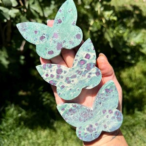 May include: Three butterfly-shaped carvings crafted from green stone with distinct magenta and deep red mineral inclusions. Each piece features a smooth, polished surface with a unique mottled pattern of dark spots and veins throughout the stone.