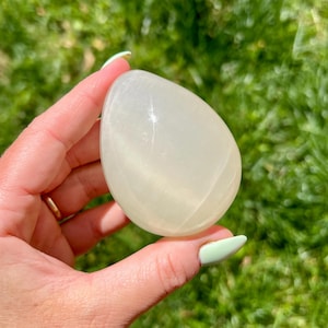 May include: A smooth, egg-shaped, off-white crystal is held in a hand against a backdrop of green grass. The crystal has a translucent quality, catching the light and reflecting a soft glow. The shape is symmetrical and polished.