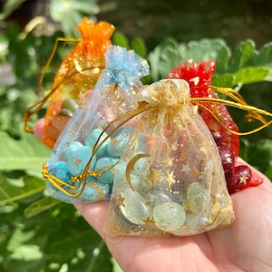 May include: A hand holds four small, sheer drawstring bags in orange, blue, gold, and red. Each bag is filled with small, polished stones. The gold bag has a star and moon pattern. The bags are tied with gold cords.