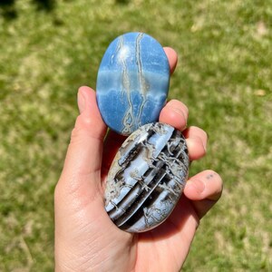 May include: Two oval-shaped polished stones. One is a light blue with white and gray veining. The other is black, white, and gray with a striped pattern. The stones are held in a hand.