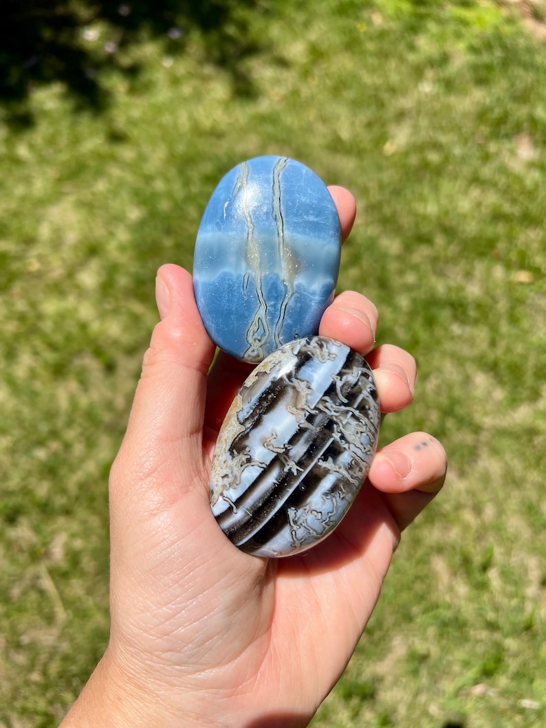 May include: Two oval-shaped polished stones. One is a light blue with white and gray veining. The other is black, white, and gray with a striped pattern. The stones are held in a hand.