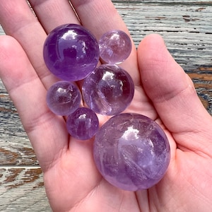 May include: A collection of six amethyst spheres of varying sizes, ranging from small to large, displayed in a hand. The spheres are a light purple color and have a smooth, polished surface.