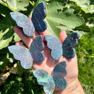 May include: Five green and pink butterfly-shaped stones with a marbled pattern. The stones are held in a hand.