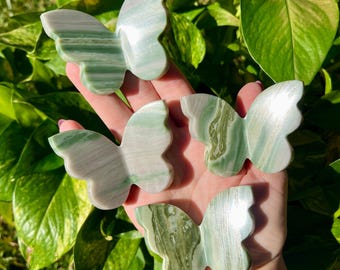 May include: Four butterfly-shaped decorative objects. Each butterfly is carved from a stone with a green and white striped pattern. The butterflies are held in a hand, with green foliage in the background.