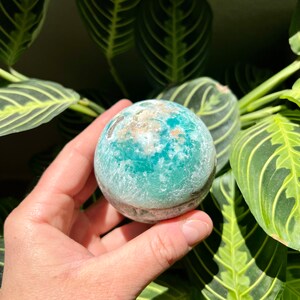 May include: A teal green and white crystal sphere with a rough, textured surface. The sphere is being held in a hand.
