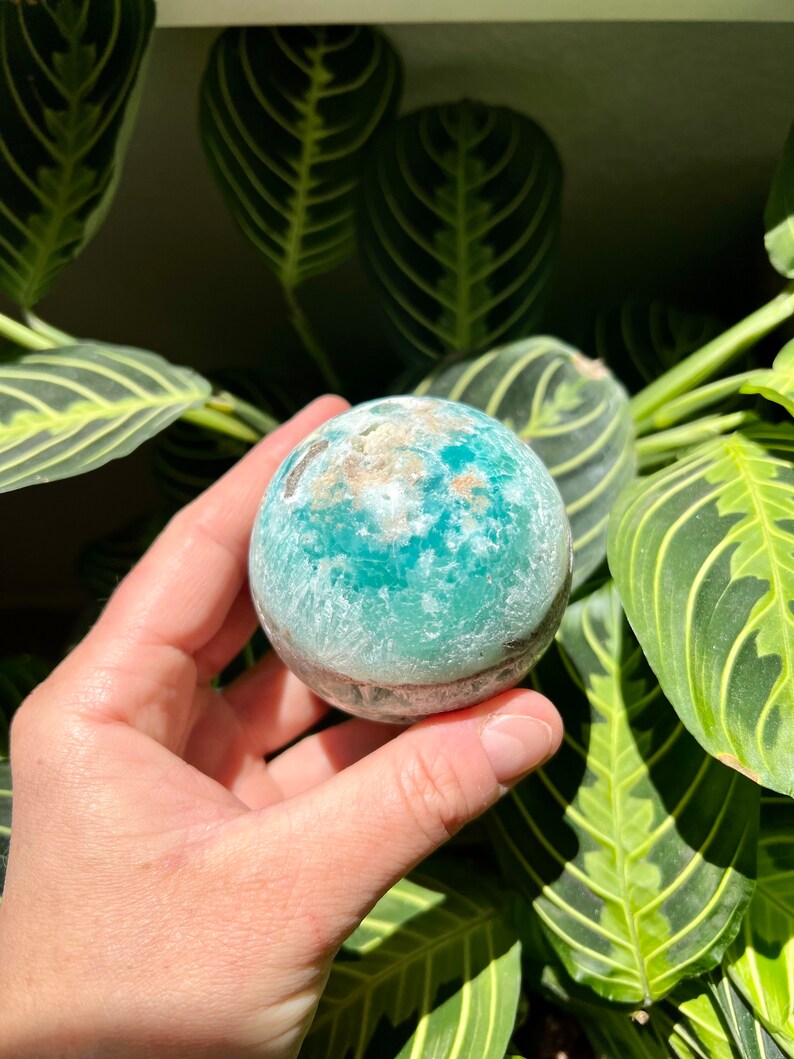 May include: A teal green and white crystal sphere with a rough, textured surface. The sphere is being held in a hand.