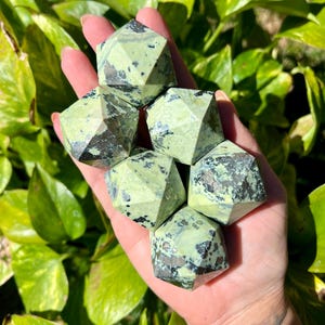 May include: Six faceted, light green gemstone dice with dark green and black inclusions are held in a hand. The dice have a geometric design and are displayed against a backdrop of green foliage.