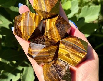 May include: A collection of five faceted tiger's eye gemstones, each featuring a distinct golden-brown and dark brown striped pattern. These polished stones are cut into geometric shapes with multiple triangular faces that reflect light.