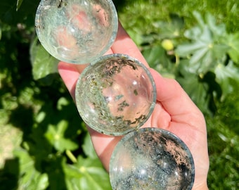 May include: Three clear, round crystal bowls with green and black inclusions are held in a hand. The bowls are displayed against a backdrop of green foliage, with sunlight illuminating the crystals.