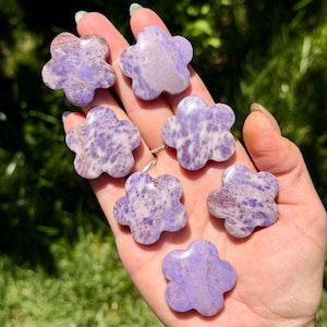May include: Seven small, flower-shaped carvings made of purple and white mottled stone. Each piece features five rounded petals and a smooth, polished surface with unique color variations ranging from light lavender to deep violet hues.