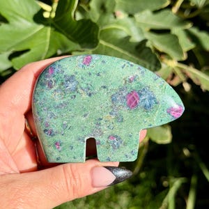 May include: A polished stone carving in the shape of a bear, featuring a light green base color with prominent inclusions of deep magenta-red and dark blue-grey mineral deposits throughout the smooth, flat surface of the object.
